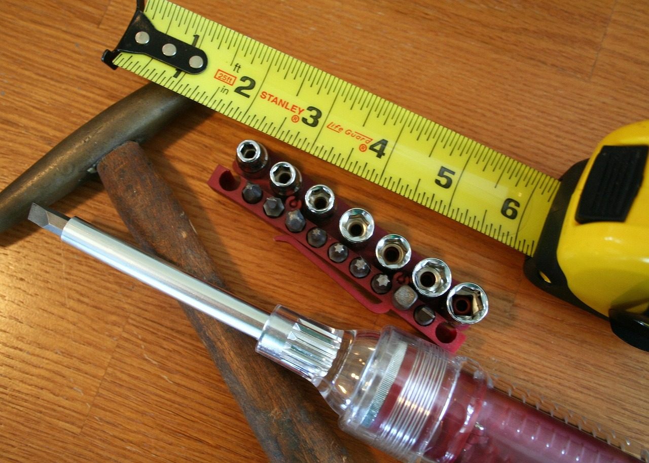 This image shows the typical tools a handyman might use in his projects such as a hammer, screw driver, sockets and a tape measure on a wood background.